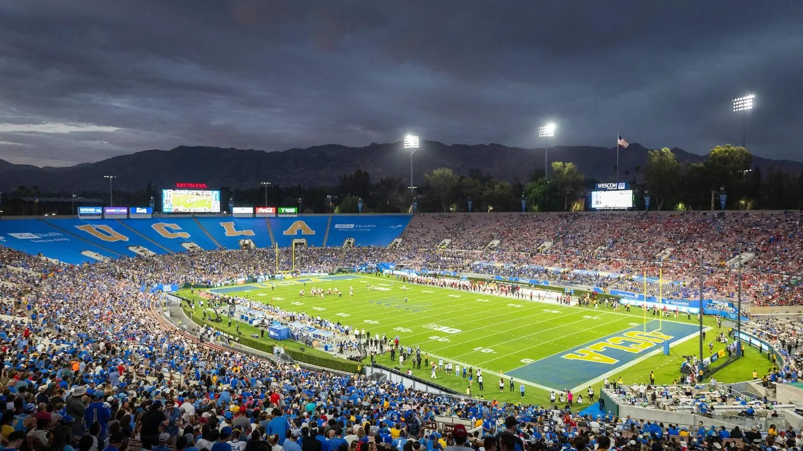 Wide view of the illuminated Rose Bowl field, packed stands and mountains beneath a darkening sky